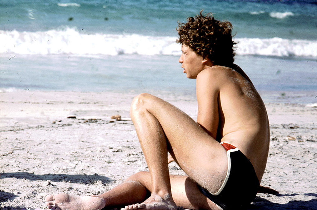 Ragazzo riccio in costume su spiaggia con bella sabbia bianca